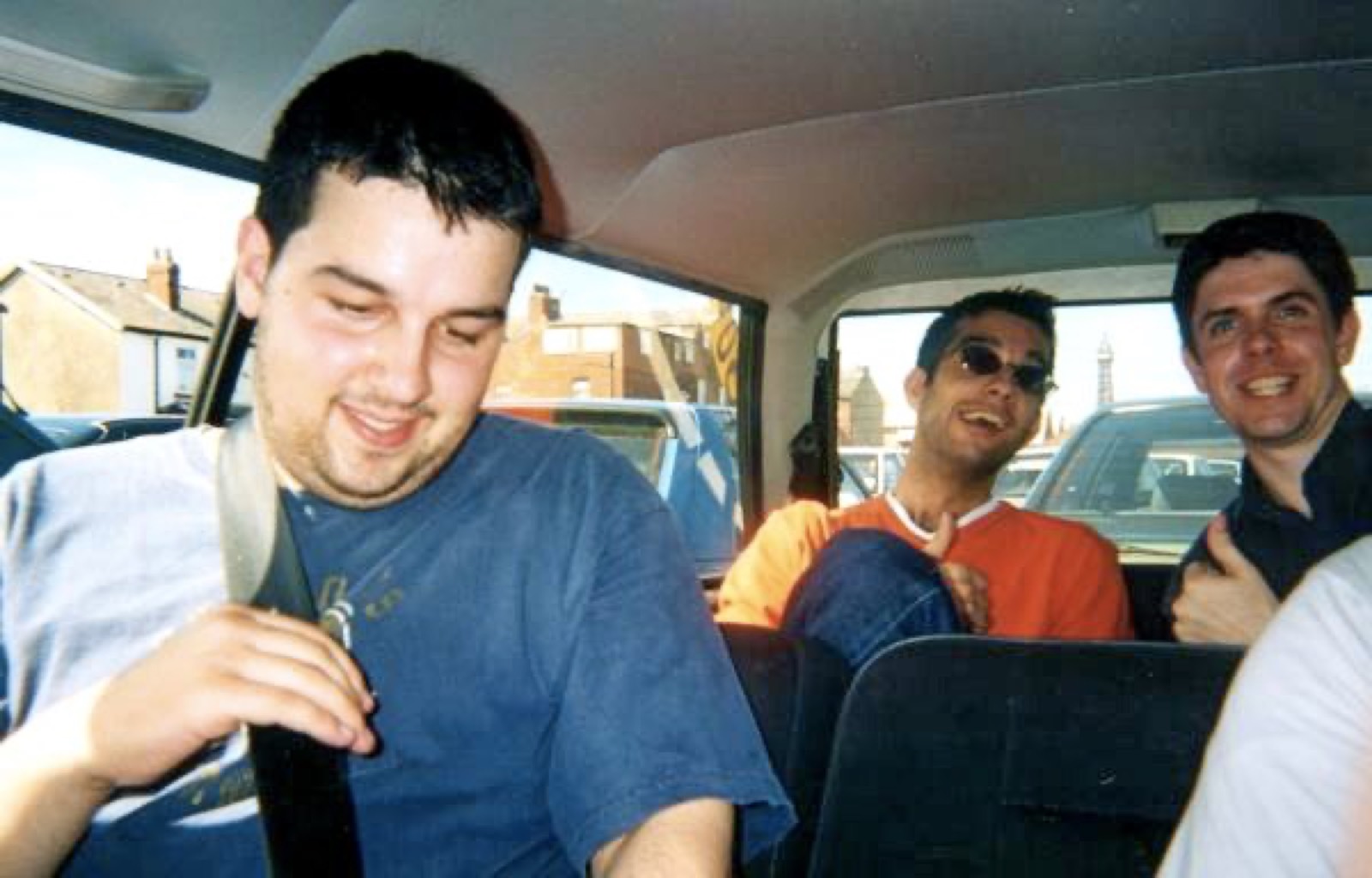 Three men laughing in the back seat of a car, one in sunglasses, terraced houses through the window behind them.