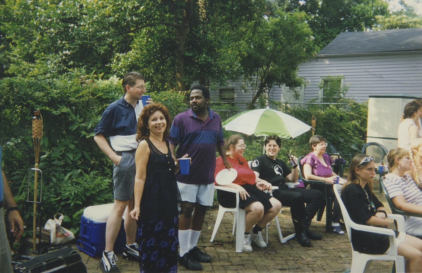 Three friends standing with blue cups in a brick back yard, a striped umbrella and more guests behind them.