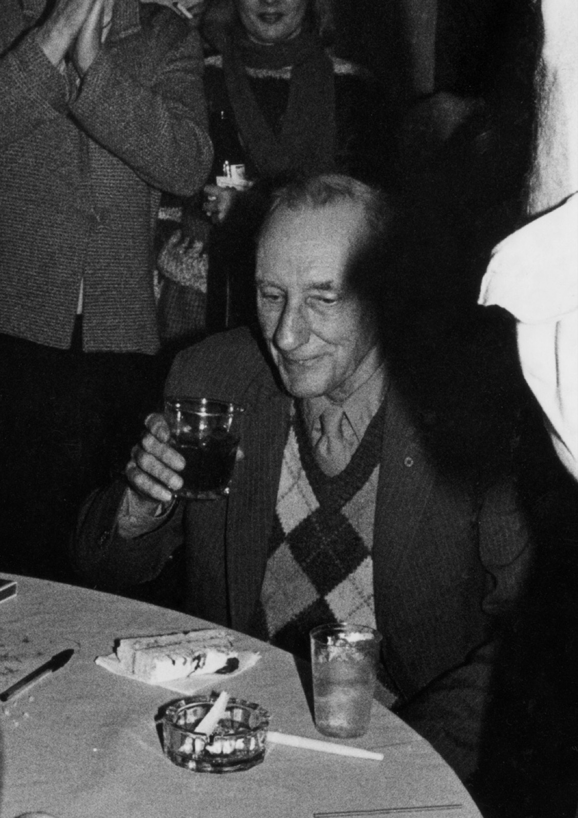 A black and white photograph of an older man at a table raising a glass, a plate of cake and an ashtray in front of him.