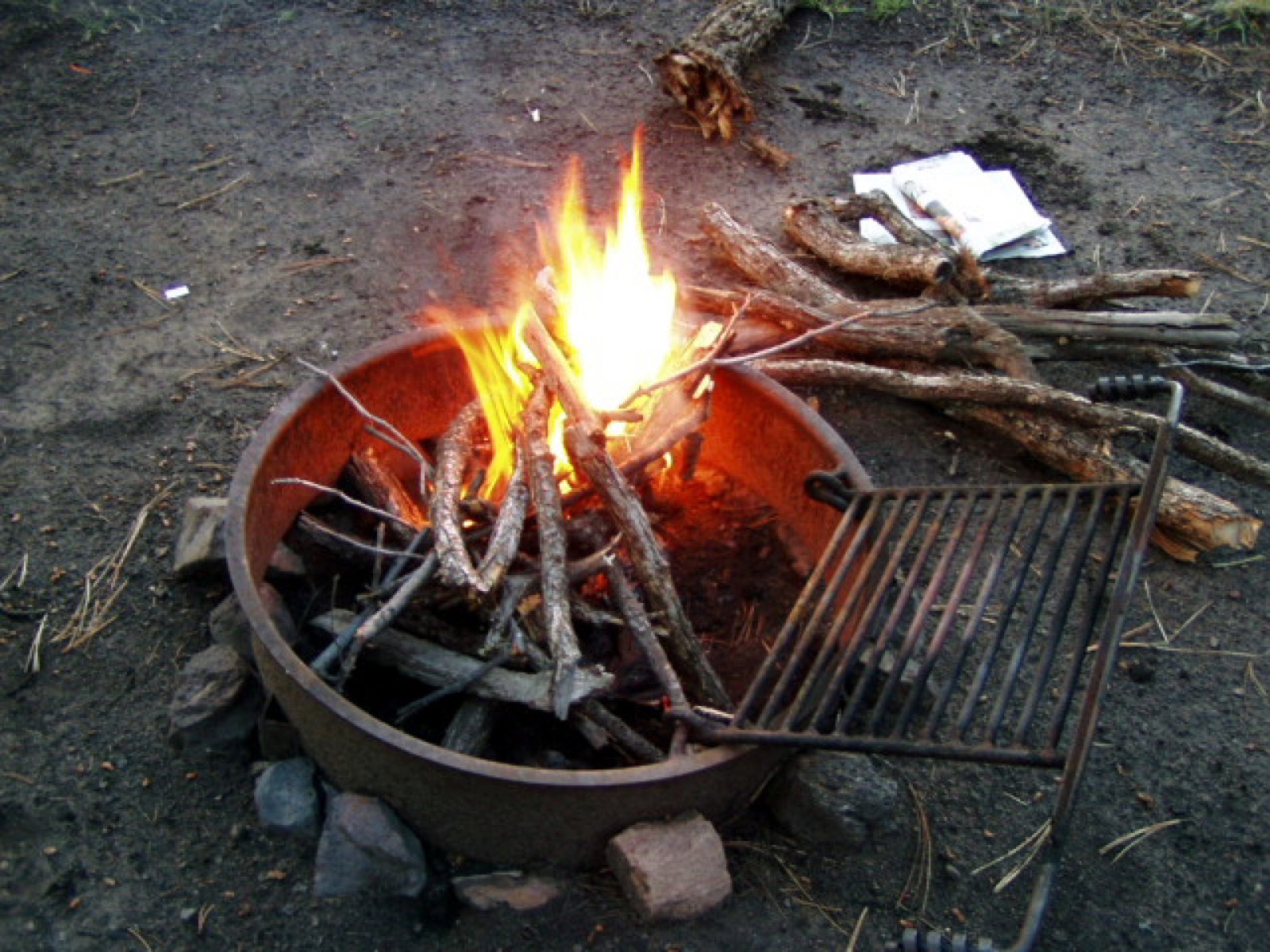 A campfire just caught in a rusted fire ring, a grate swung to one side and a pile of kindling beside it.