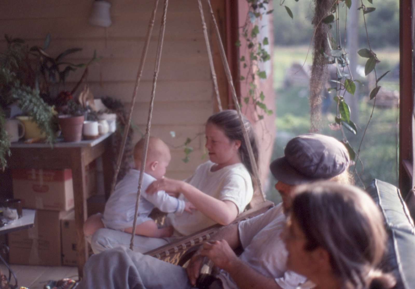 A young woman holding a baby in a hanging rope chair on a front porch, potted plants behind her, a man in a cap beside them.