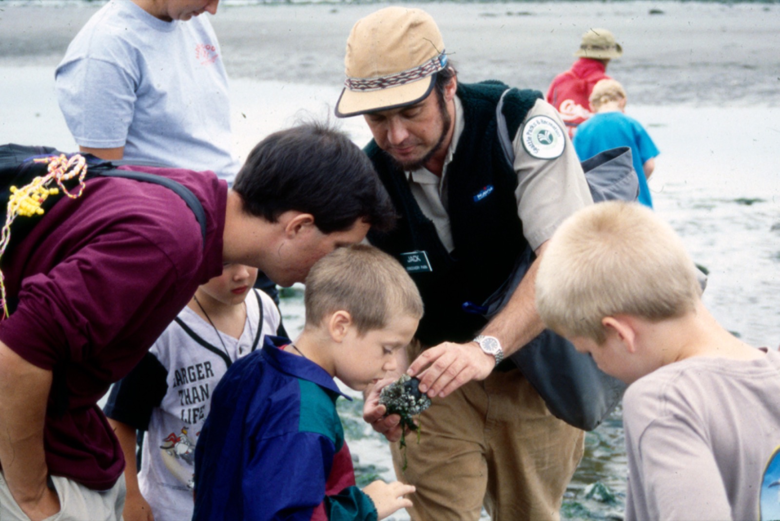 A park ranger holding a rock covered in barnacles while a ring of children lean in to look.