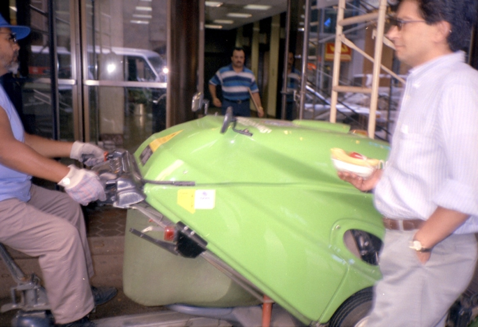 A street sweeper's cart, bright green, stopped on a sidewalk while a man with a hot dog looks on.
