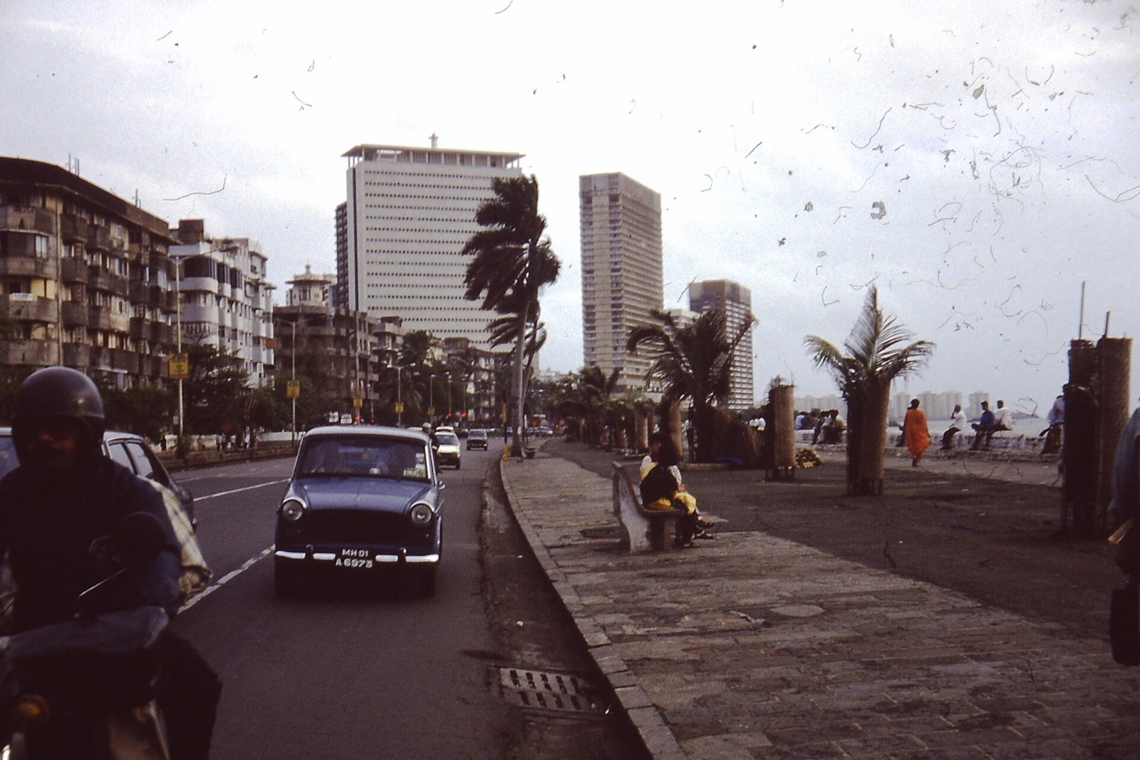 A wide seafront road with a blue Ambassador car, a scooter rider in a helmet, palms and towers beyond.