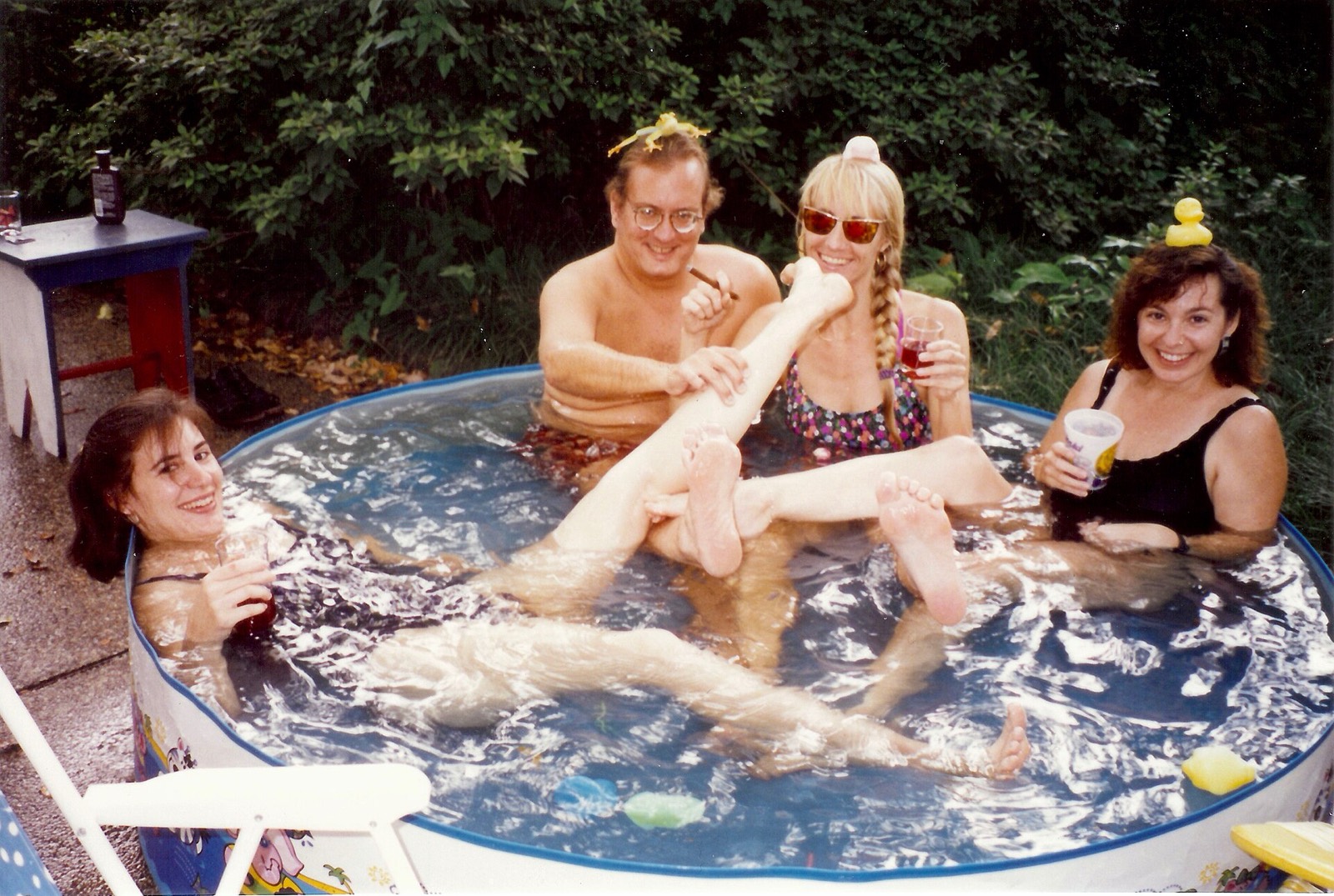 Four friends squeezed into a kiddie pool, legs everywhere, rubber ducks balanced on their heads.