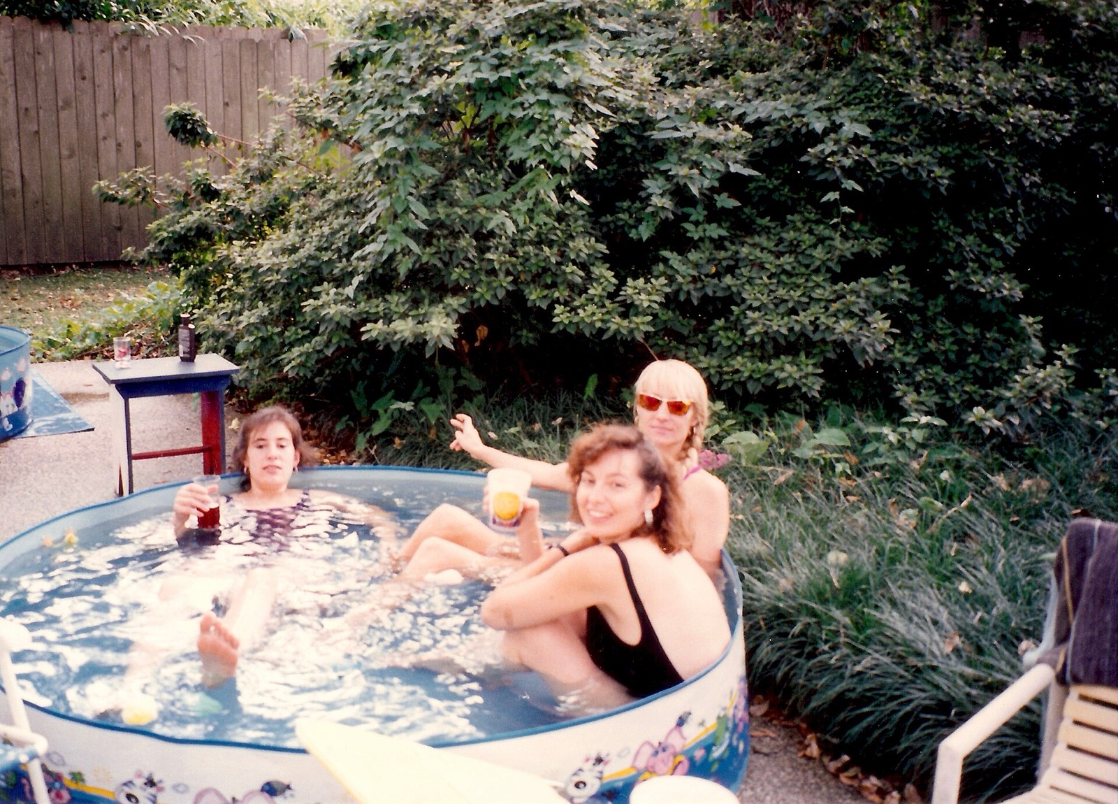 Three women with drinks in an inflatable kiddie pool at the edge of a garden, one waving at the camera.