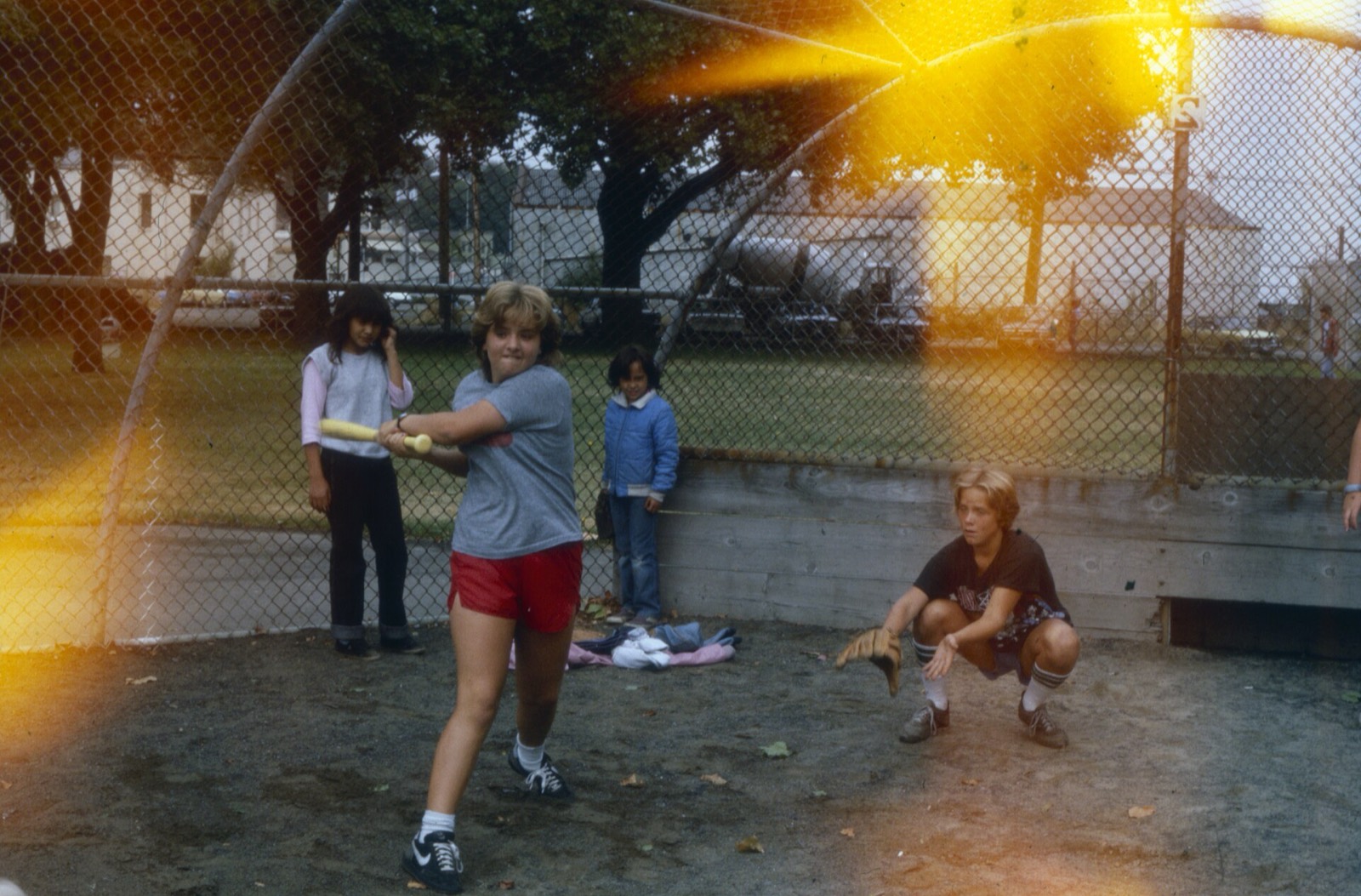 A girl in red shorts mid-swing at a backstop, a catcher crouched behind her, sunlight flaring through the fence.