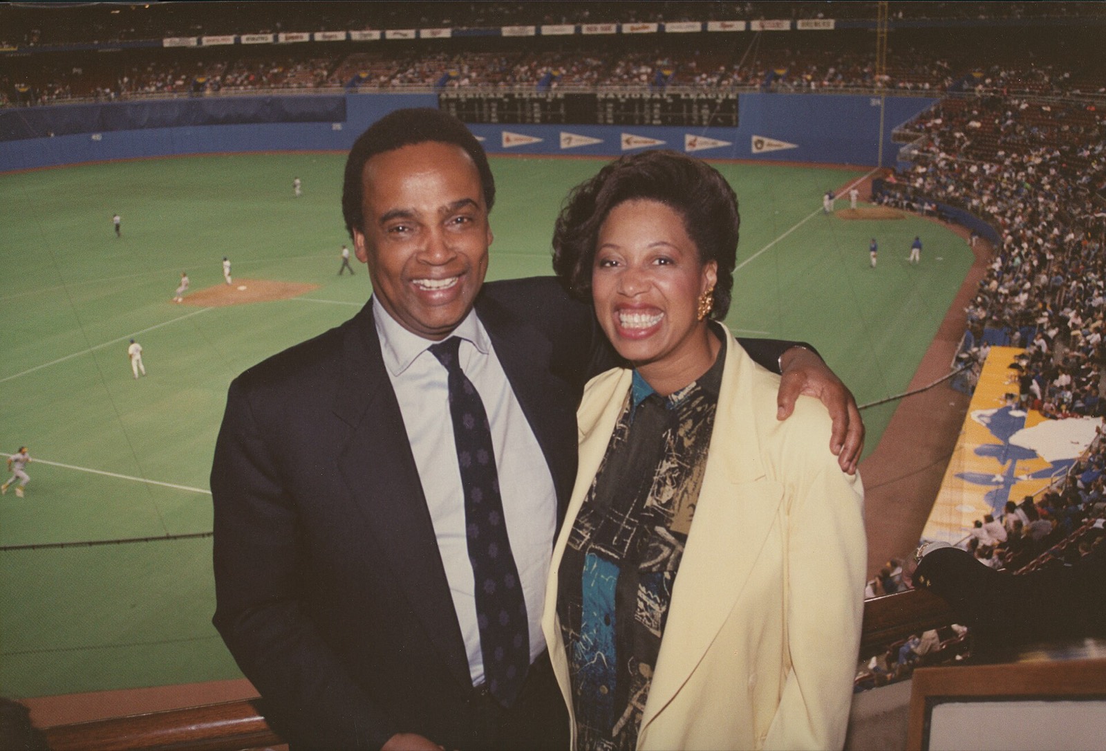 A couple in evening clothes laughing arm in arm above a lit baseball diamond.