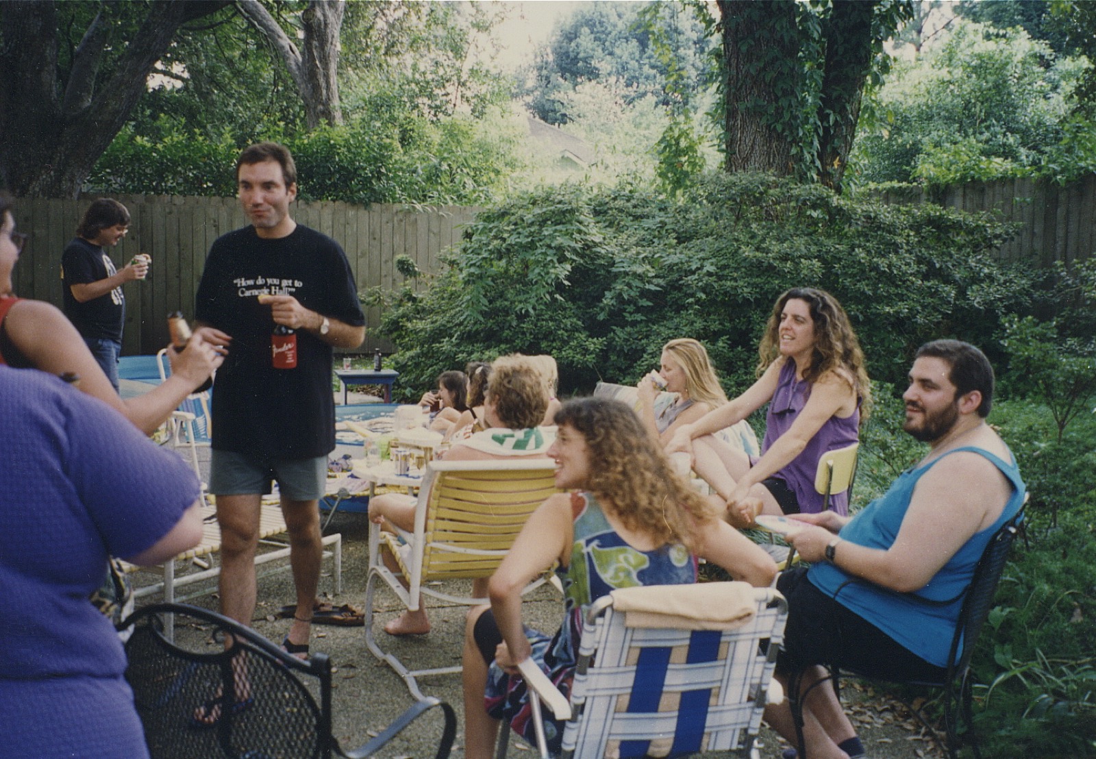 Friends in lawn chairs in a green back yard, one standing with a drink, the others turned toward each other.