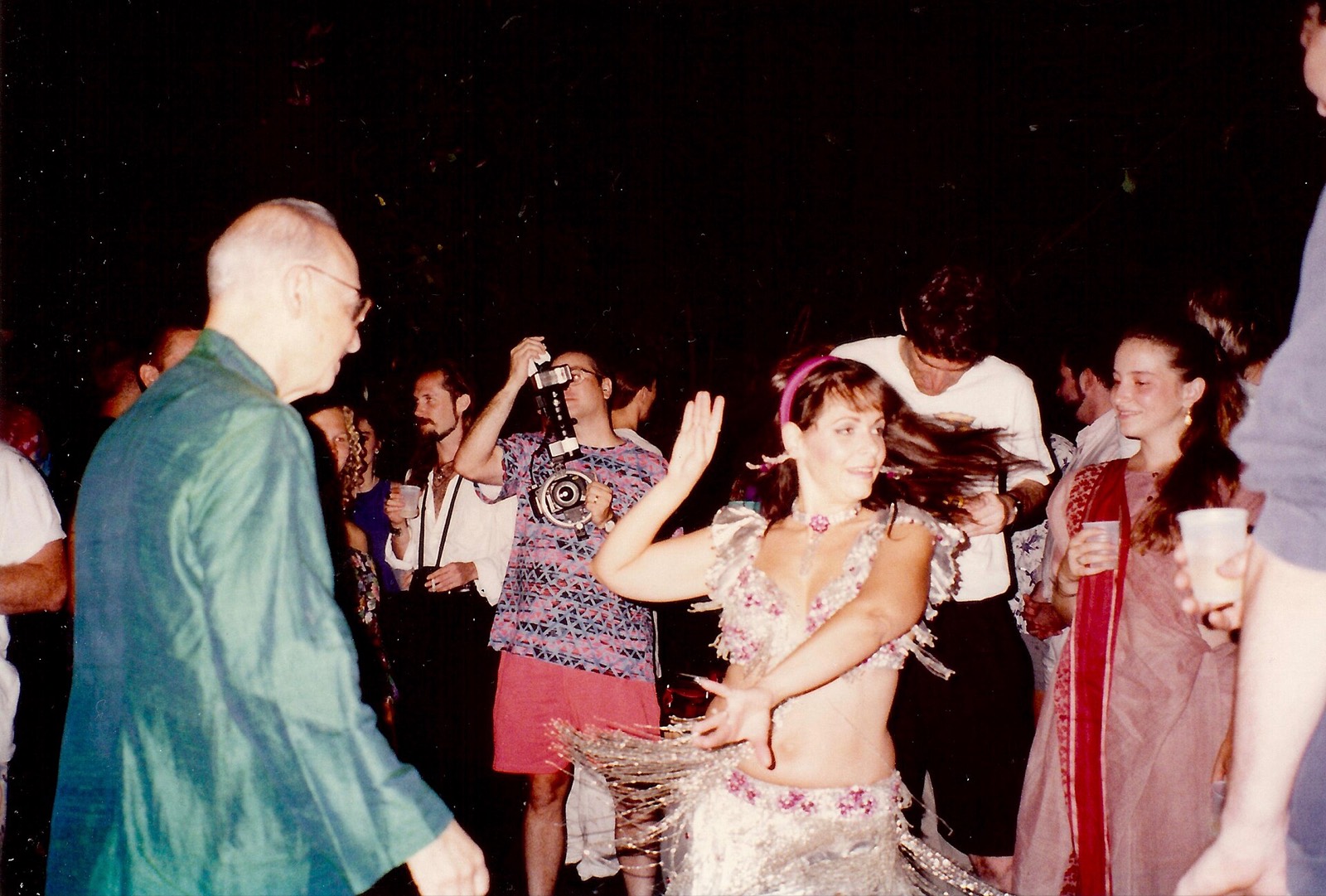 A night party: a dancer in sequins, a man in a green shirt, someone holding a camera up over the crowd.