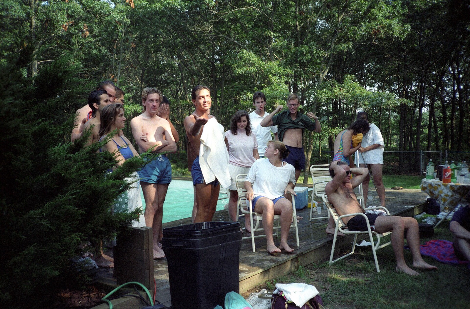 A crowd of friends on a pool deck in the trees, one of them mid-sentence, watching a band play.