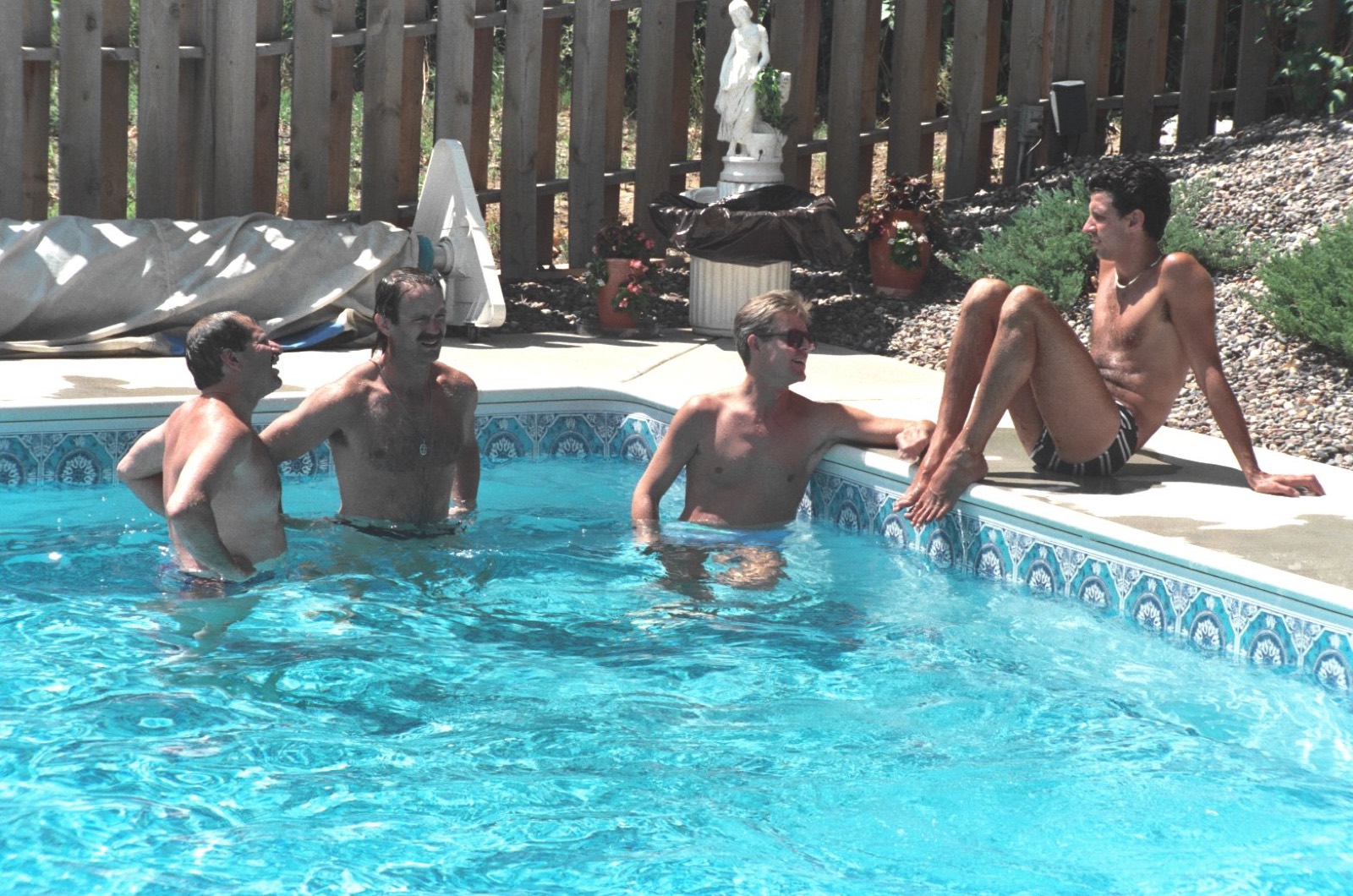 Three men talking in a backyard pool while a fourth sits on the edge with his knees up.