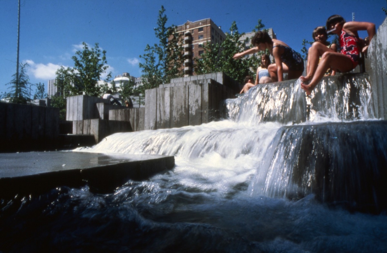 Children in swimsuits perched on the concrete ledges of a park fountain as water pours over the steps below them.