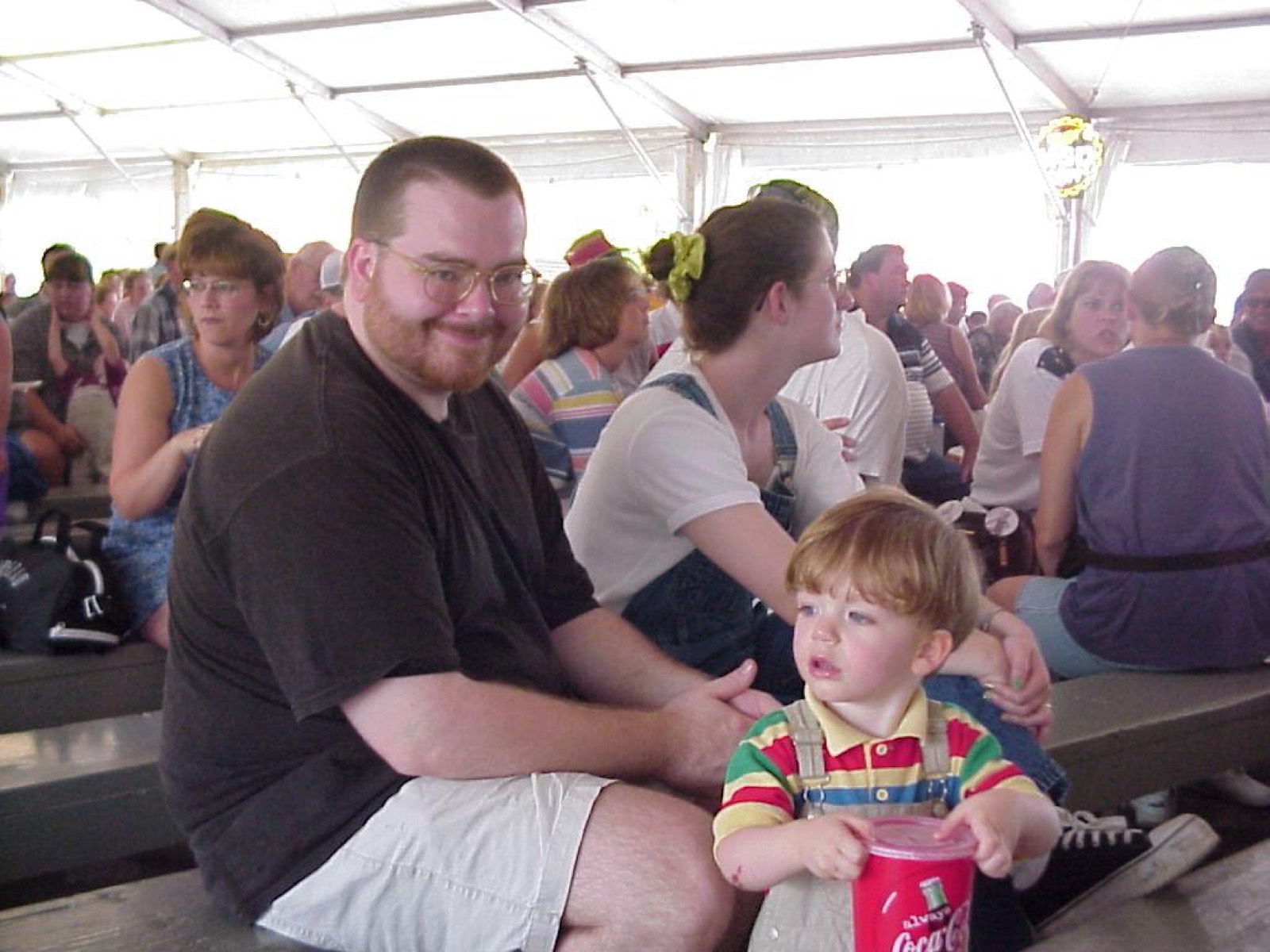 A man in glasses smiling on a bleacher under a white tent, a toddler in a striped shirt clutching a cup in front of him.