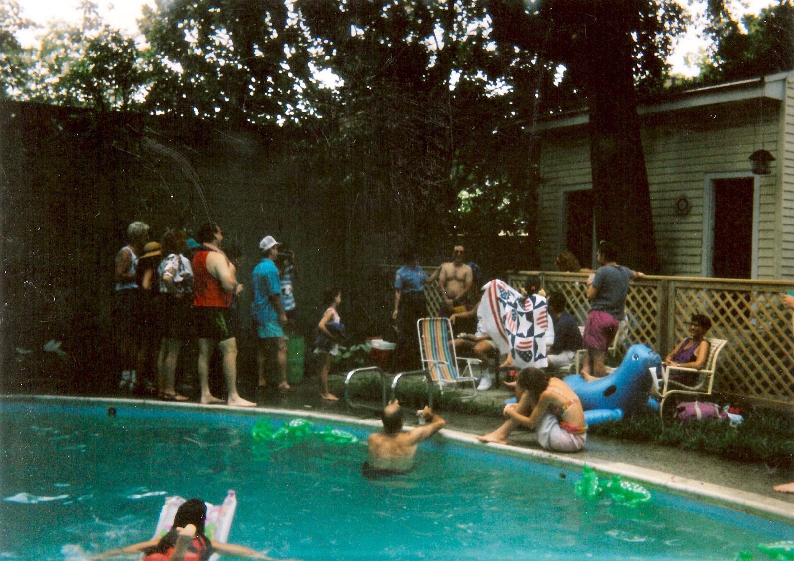 A shaded backyard pool with people standing along the fence, a striped chair, a blue inflatable dinosaur.