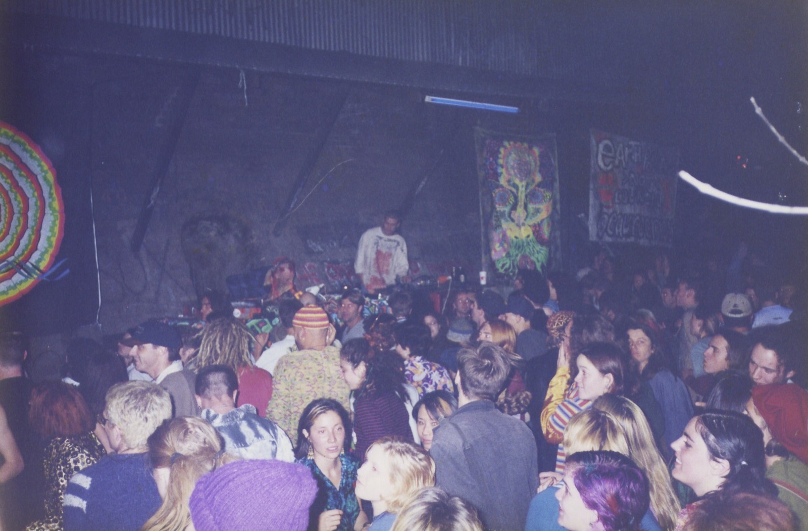 A dark warehouse crowd turned toward a DJ under hand-painted banners, faces lit from one side.
