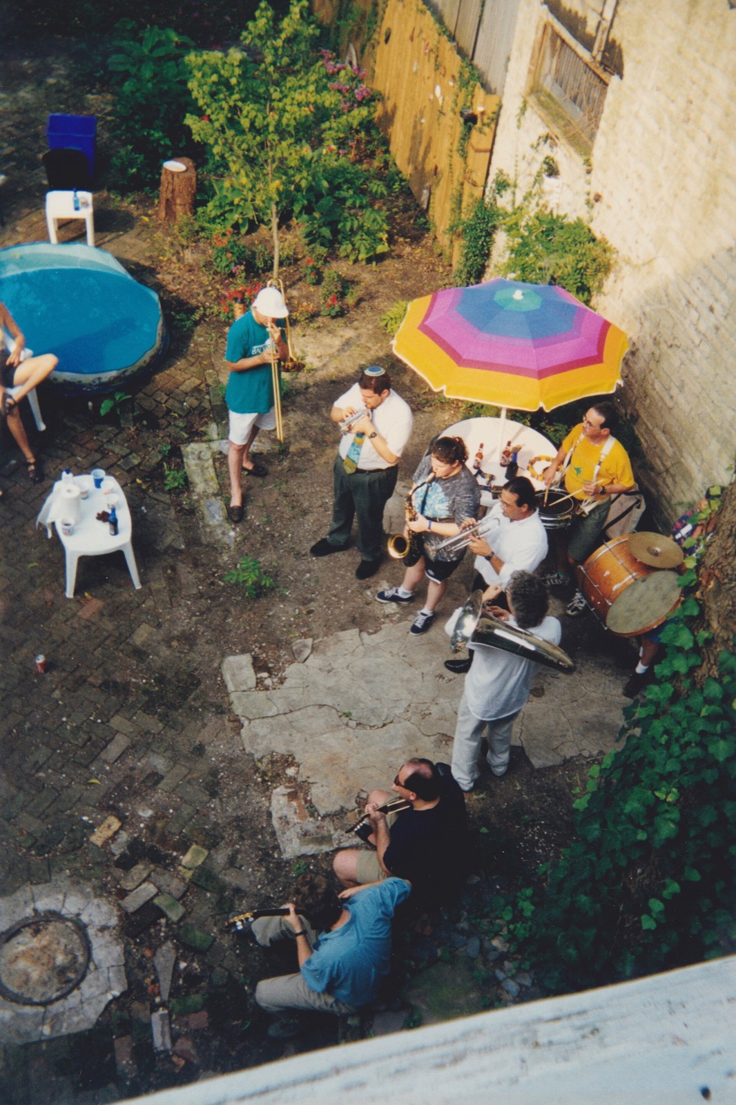 A backyard party seen from an upstairs window: a small brass band under a striped umbrella, a wading pool, plastic chairs on brick.