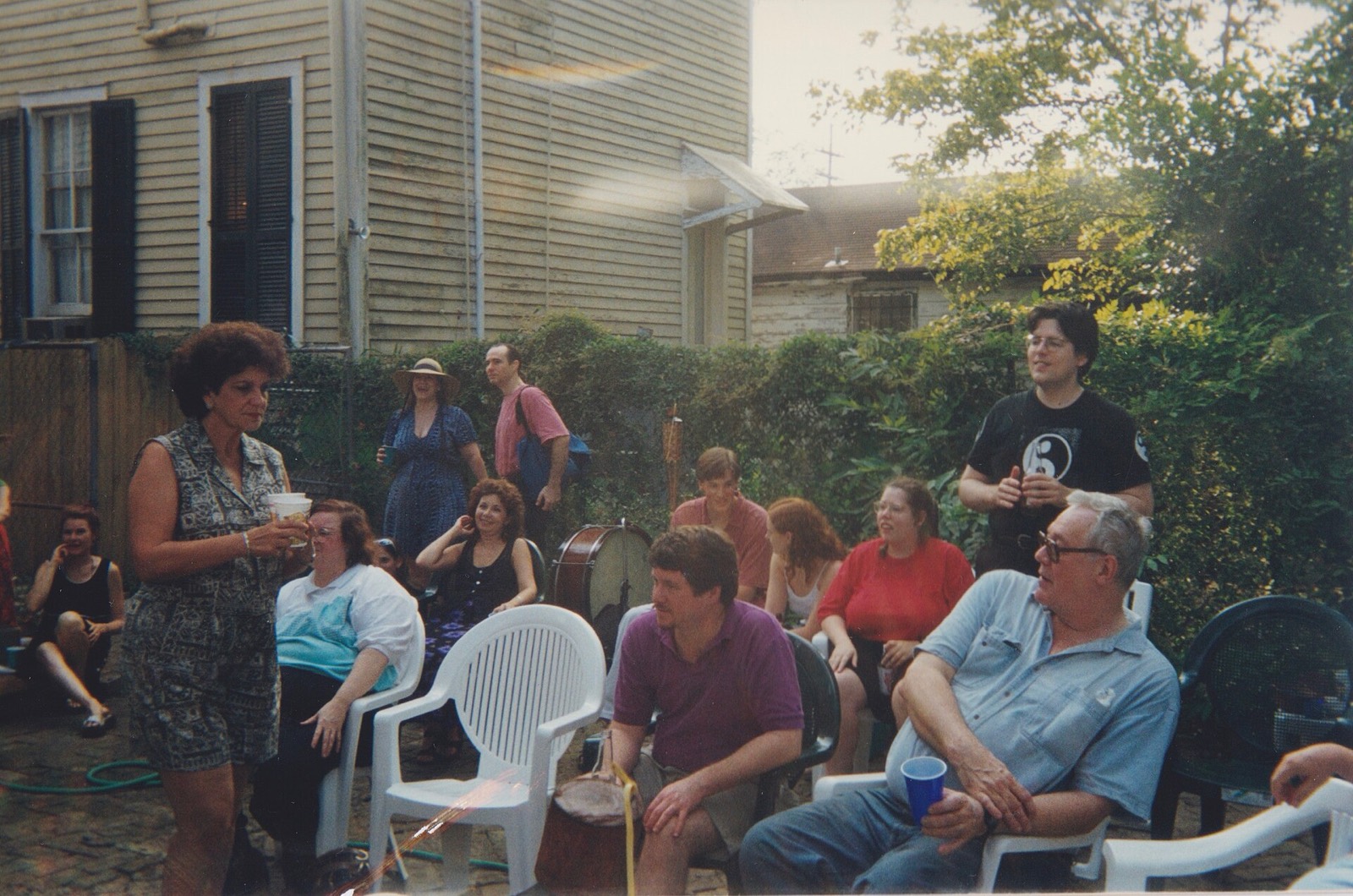 A backyard party in plastic chairs beside a clapboard house, a woman walking through with a cup.