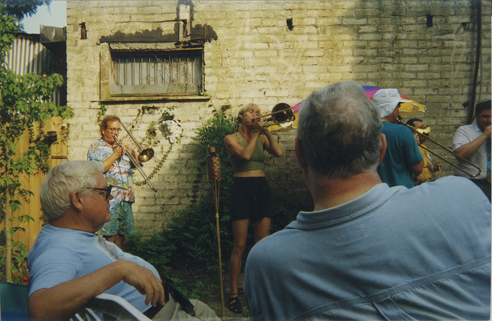 Three trombonists playing against a whitewashed brick wall while two men listen from lawn chairs.
