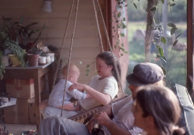 A young woman holding a baby in a hanging rope chair on a front porch, potted plants behind her, a man in a cap beside them.