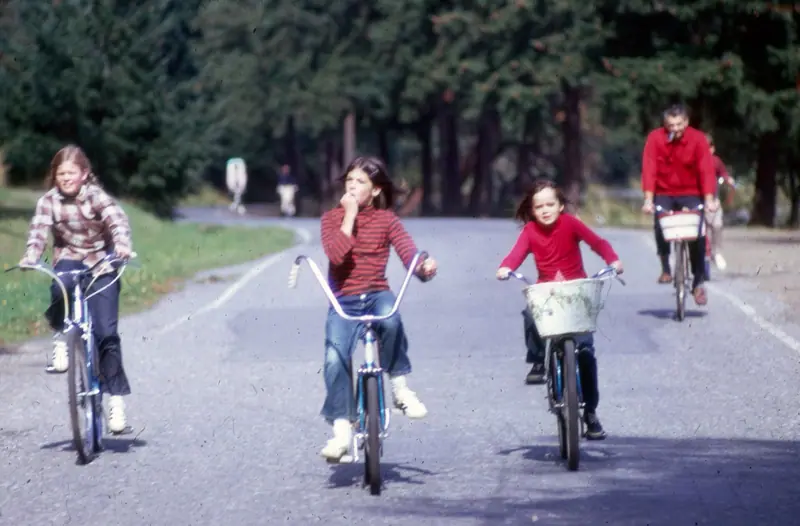 Three girls and a man riding bicycles up a park road under tall firs, one girl with a white basket.