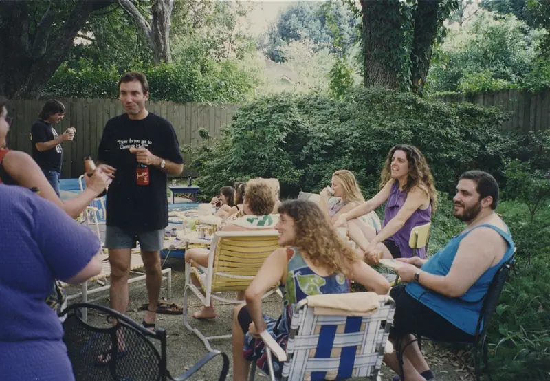 Friends in lawn chairs in a green back yard, one standing with a drink, the others turned toward each other.